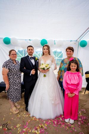 Wedding celebration with guests in formal wear gathered under a white tent with teal balloon decorations.