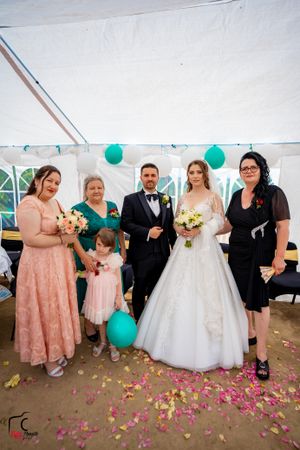 Wedding group photo with guests wearing pink dresses and teal accents under a white tent.