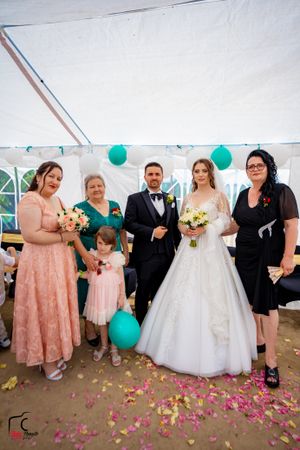 Wedding party members gather under a tent with teal balloons and scattered pink petals on the ground.