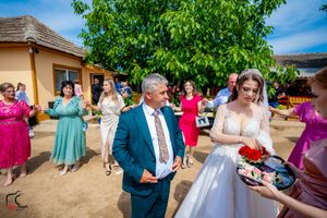 Wedding celebration outdoors with guests in colorful dresses dancing near a beige building under a green tree.