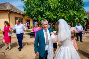 Wedding party dancing outdoors near a tan building with guests in bright formal attire.