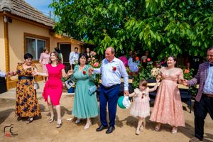 Wedding party dances in a line outside a rustic venue with green trees in background.
