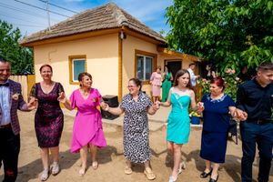 Celebratory group performing a traditional dance line outside a rustic yellow house.