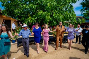 Colorfully dressed guests performing a group dance at an outdoor celebration under leafy trees.