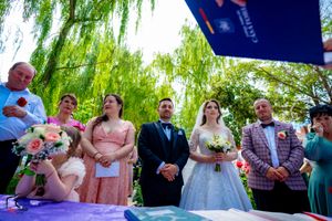 Wedding guests watch ceremony proceedings at a purple-decorated table beneath lush willow trees.