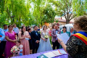 Guests gather for an outdoor wedding ceremony beneath green willow trees with purple table decorations.