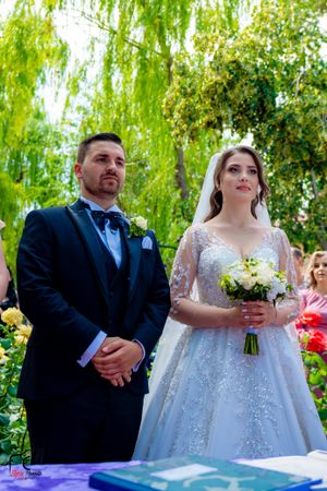 Wedding couple stands at ceremony table with white floral bouquet under willow trees.