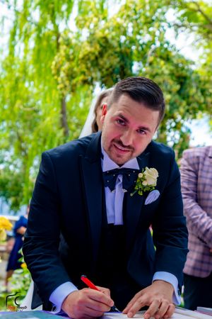 Groom in black tuxedo signs documents at outdoor wedding ceremony.