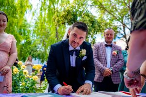 Wedding guests gather under a willow tree to sign documents at an outdoor ceremony.