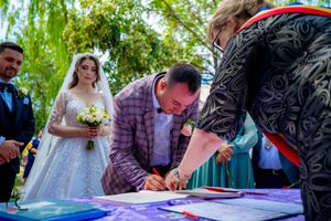 Wedding party members gathered around table for document signing under green trees.