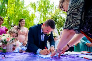 Wedding ceremony guests gather outdoors under a willow tree to sign official documents on a purple table.