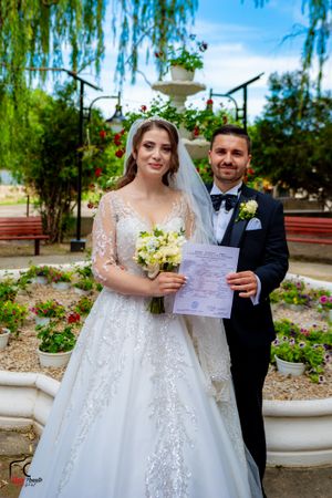 Newlyweds holding documents and bouquet in garden setting with willow trees.