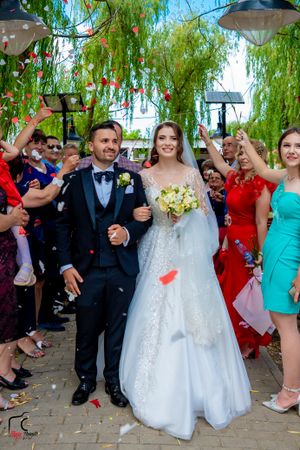 Wedding party cheers and throws petals during outdoor celebration under weeping willow trees.