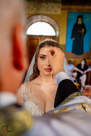 A bride wearing a sparkling dress and veil has a red bindi applied to her forehead during a ceremony.