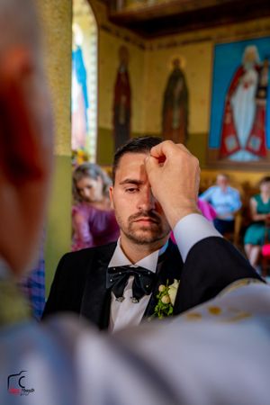 A groom in a black tuxedo receives a red mark on his forehead during a traditional wedding ceremony.