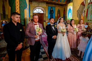 Wedding party and guests stand in an Orthodox church with colorful religious murals and golden columns.