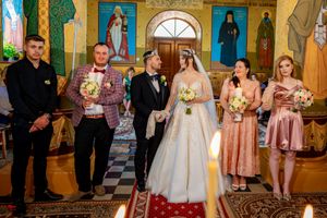 Wedding ceremony participants arranged in an Orthodox church with colorful religious murals and golden accents.