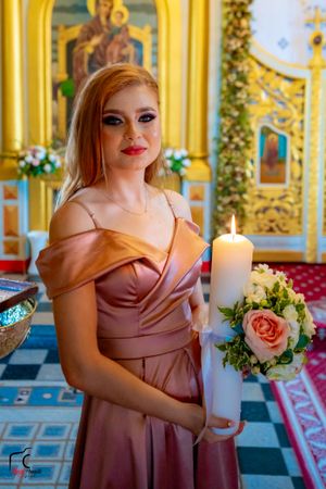 Bridesmaid in dusty rose dress holding ceremonial candle with floral arrangement in Orthodox church.