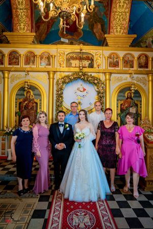 Wedding ceremony group gathered in Orthodox church with elaborate gold iconostasis and black and white tile floor.