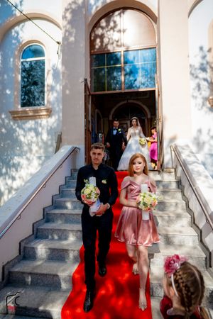 Wedding party exits church onto red carpet with white doves in foreground at outdoor ceremony.