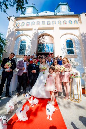 Wedding party and guests pose outside church with white doves on red carpet.