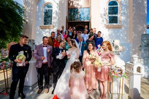 Wedding party poses together outside a church with white walls and arched windows.