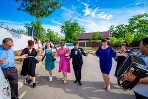A festive wedding party dances in a line outside on a sunny day with bright blue skies and green trees.