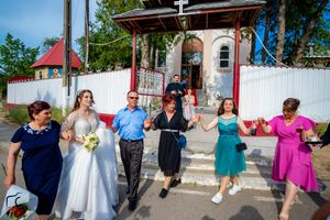 A wedding party performs a traditional line dance outside a historic church building with white fencing.