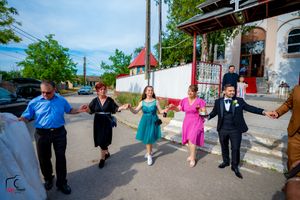 Wedding guests perform a celebratory dance line outside a white-fenced church on a sunny afternoon.