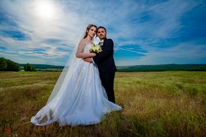 Newlyweds pose together in a scenic field under bright blue skies with rolling hills in background.