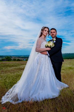 Elegant wedding couple embraces in field with flowing white dress and dramatic sky backdrop.