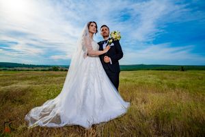 Newlyweds stand together in an open field with dramatic clouds and natural landscape.