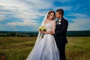 Bride in flowing white gown and groom share intimate moment in rural meadow setting.