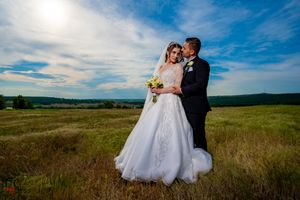 A wedding couple poses in a scenic meadow with rolling hills and blue skies in the background.