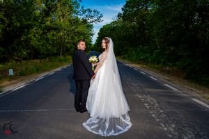 Wedding couple stands together on an empty rural road surrounded by lush green trees.