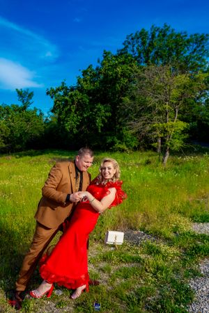 Couple in brown suit and red dress pose playfully in a sunny meadow.