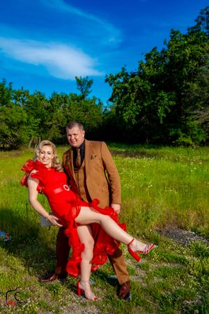 Playful couple in formal wear poses dramatically in a sunlit field.