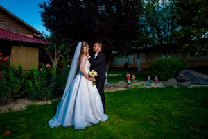 A romantic wedding couple poses on a lush garden lawn at dusk with a rustic building in the background.
