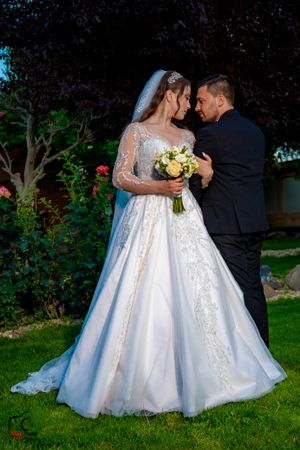 An elegant wedding couple embraces while holding a yellow bouquet in a landscaped garden.