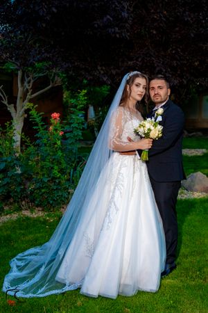 Bride in lace gown and groom in dark suit embrace in garden setting at sunset.