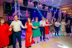 People in bright formal wear dancing in a line at an indoor event with stage lighting.