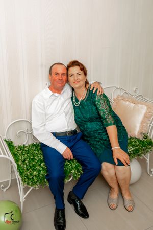 Couple poses on decorative white bench with greenery and floral arrangements in background.