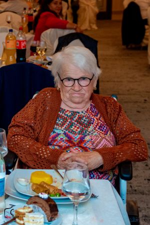An elderly person in a brown sweater sits at a dining table with food and drinks.