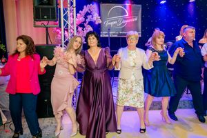 Party guests in formal attire dance in formation during an indoor celebration with colorful lighting.