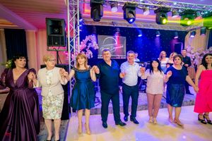 Celebrants in formal wear participate in a group dance under vibrant stage lighting at an indoor venue.