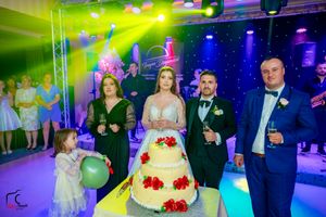 A wedding party gathers around a beautiful tiered cake with red roses under vibrant lighting effects.