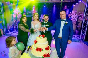 Wedding party poses beside a multi-tiered white cake with red rose decorations under colorful spotlights.