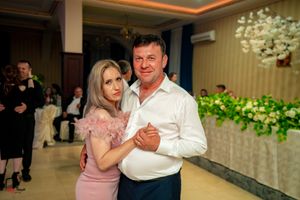 Couple dancing together at an elegant event with pink dress and white shirt against floral backdrop.