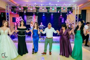Formal party guests hold hands in a dance line under vibrant stage lighting at an indoor celebration.