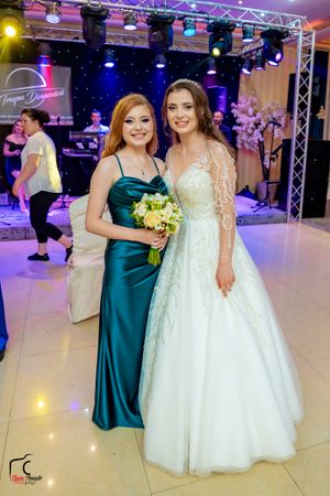 Two attendees pose at a wedding reception with stage lighting and decorative floral arrangements.